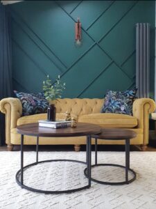 The image showcases a vibrant living space with a yellow chesterfield sofa against a bold green geometric wall, creating a striking contrast. Tropical-patterned pillows enhance the color scheme, while two modern coffee tables with wooden tops and metal bases add style and functionality. A potted plant on the table brings a touch of greenery, complemented by decorative books and accents. A pendant light provides illumination, and tall windows with curtains allow natural light to fill the room. A great example of an Interior design for people who want to go From Beige to Bold