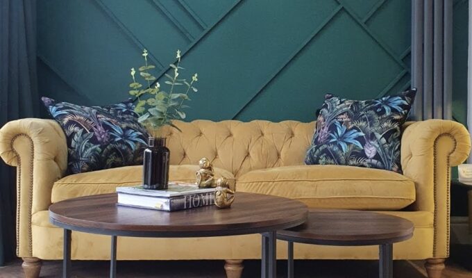 The image showcases a vibrant living space with a yellow chesterfield sofa against a bold green geometric wall, creating a striking contrast. Tropical-patterned pillows enhance the color scheme, while two modern coffee tables with wooden tops and metal bases add style and functionality. A potted plant on the table brings a touch of greenery, complemented by decorative books and accents. A pendant light provides illumination, and tall windows with curtains allow natural light to fill the room. A great example of an Interior design for people who want to go From Beige to Bold