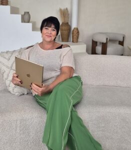 A woman with short dark hair sits comfortably on a textured beige sofa, holding a gold-colored tablet. She is wearing a light beige knit top with a black strap visible underneath and loose, vibrant green pants with white piping. The background features minimalist decor, including neutral-toned vases and pampas grass arranged on a white stepped surface. A modern armchair with wooden legs and a plush seat is visible in the corner, contributing to the overall serene and stylish ambiance. She is waiting for her clients to contact her to discuss their projects.