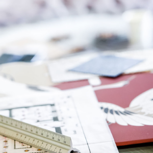 A close-up of a workspace featuring various design materials scattered on a table. A ruler is prominently placed on top of a floor plan, while nearby, fabric swatches and samples are visible, including a red piece with a white crane illustration. The items suggest an interior design or architectural project in progress, with a focus on textiles, patterns, and precise measurements